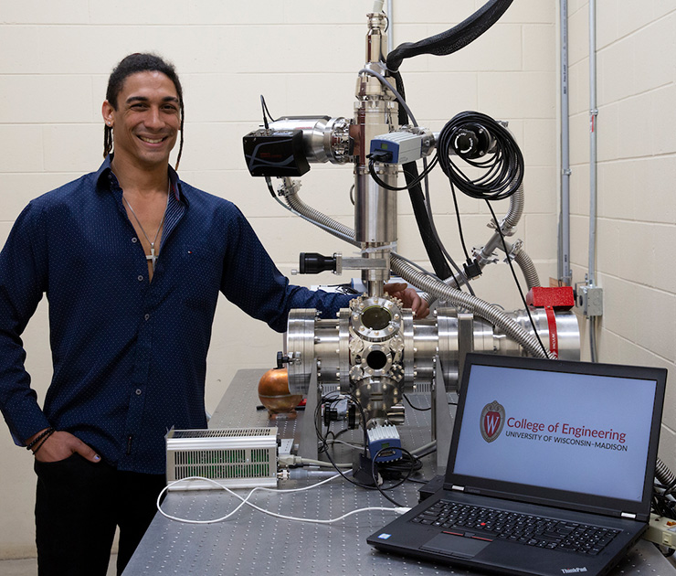 A smiling graduate student stands next to the complex-looking apparatus he developed.