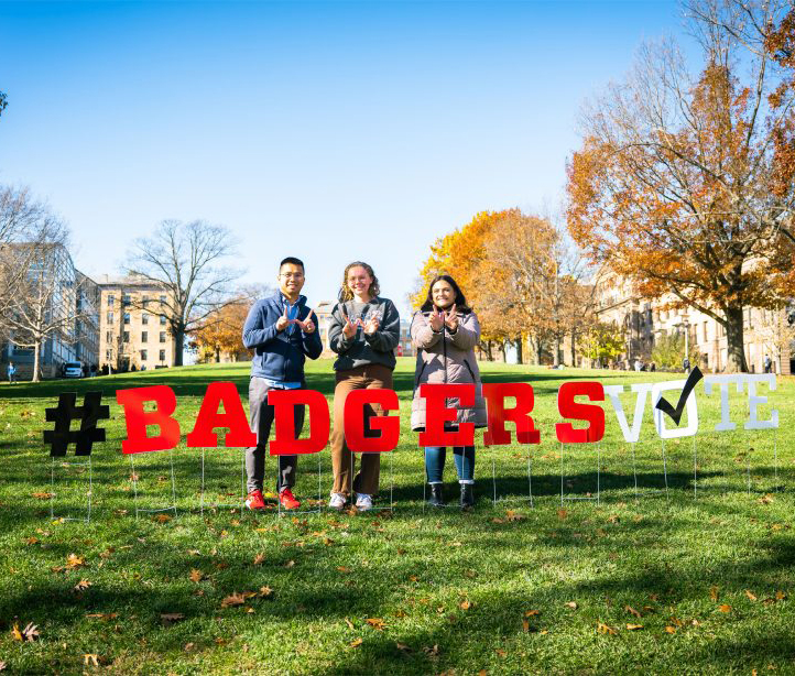 Three individuals standing on Bascom Hill, each making the W sign with their hands. They are standing behind yard signs spelling "#BADGERSVOTE".