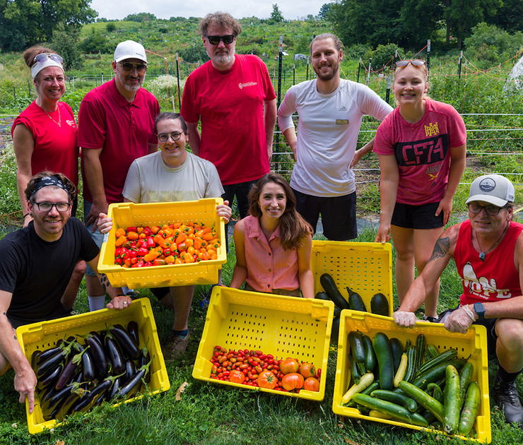 Eight people stand and display vegetables they picked, collected in large yellow bins.